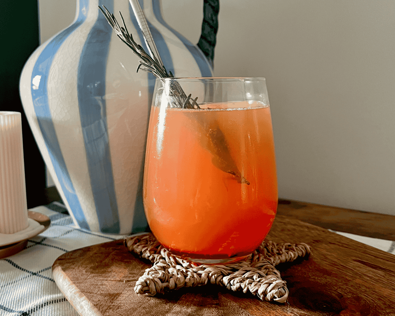A drink called the Shine Starburst is in the focal point. It's an orange drink with a sprig of rosemary added for decoration. It's sat on a star-shaped coaster in front of a white and blue decorative vase.