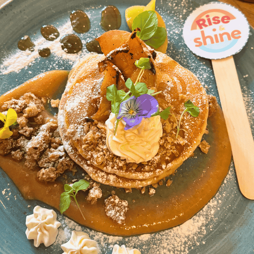 A stack of pancakes with bacon, whipped cream, and edible flowers. The Rise and Shine logo appears in the top right corner.